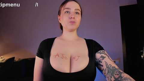 Media: Video of a light-skinned woman with dark hair, wearing a low-cut black top revealing large breasts with \"A-JE5\" tattooed on her chest. She has a sleeve tattoo on her left arm. The background is dimly lit with purple hues, and a \"rip menu\" watermark is visible.