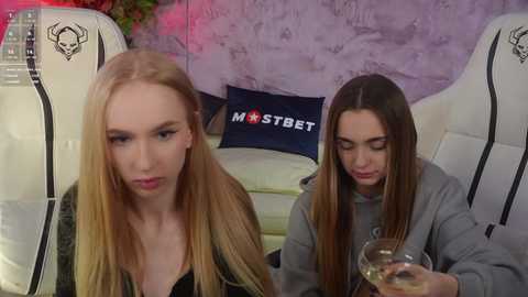 Media: Video of two young women with fair skin, long straight hair, and medium-sized breasts, sitting in a modern living room. One woman sips wine, the other looks contemplative.