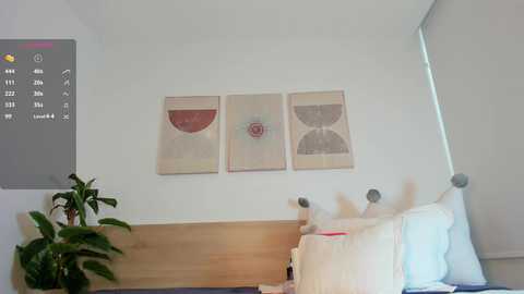 Media: A minimalist bedroom video featuring a white bed with white pillows, a wooden headboard, and a beige wall adorned with three abstract, circular, pastel-hued paintings. A small plant is on the left, and a grey digital clock is mounted on the wall.