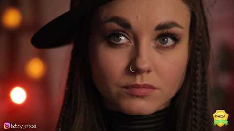 Media: Video of a woman with light brown skin, green eyes, and dark hair under a black cap, wearing a black turtleneck. Background features warm, blurred lights and a \"OWN\" logo in the corner.