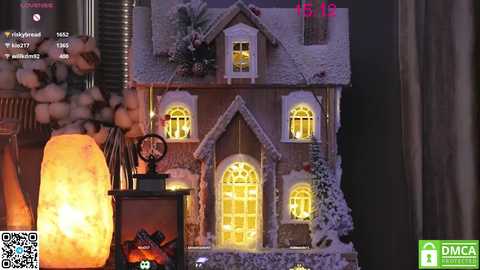 Media: Video of a small, intricately decorated, snow-covered gingerbread house with glowing windows and a lit fireplace, surrounded by candles and a cozy atmosphere.