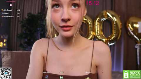 Media: Video of a young woman with light skin, blonde hair, and blue eyes, wearing a brown spaghetti strap top, surrounded by gold \"100\" balloons in a dimly lit room.