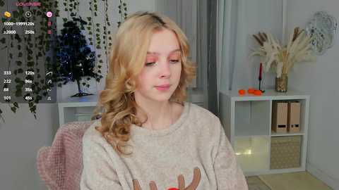 Media: A young, fair-skinned woman with wavy blonde hair sits indoors, wearing a beige sweater with cartoon reindeers. The room features a white bookshelf, greenery, and a vase with orange slices.