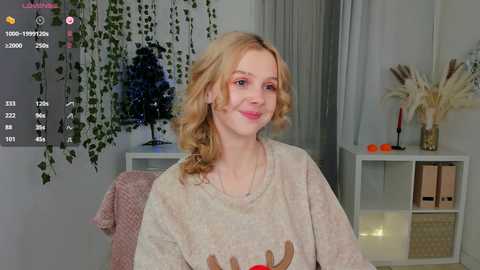 Media: Video of a smiling young woman with curly blonde hair, wearing a beige sweatshirt, in a cozy, greenery-filled living room.