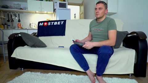 Media: Video of a Caucasian man with short hair, wearing a green t-shirt and blue jeans, sitting on a cream couch with a \"MOSTBET\" towel draped over the back, in a modern, minimalist kitchen.