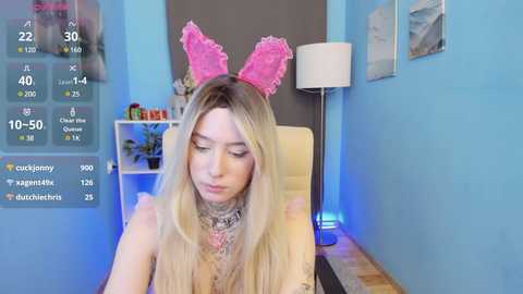 Media: Video of a young, fair-skinned woman with long blonde hair wearing a pink bunny headband, sitting at a desk in a light blue room with a white lamp.