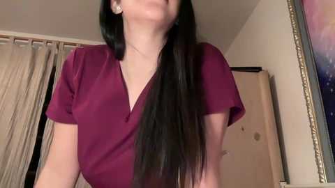 Media: Video of a woman with long black hair, wearing a deep purple V-neck shirt, standing indoors. Background features beige curtains, a white door, and a partially visible colorful painting.