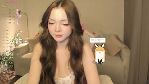 Media: Video of a young Asian woman with long, wavy brown hair, wearing a white lace top, sitting in a cozy living room with beige furniture and a fox sticker on her shirt.