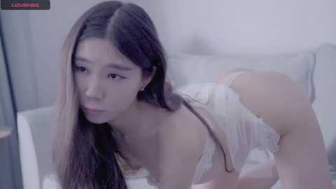 Media: Video of an East Asian woman with long black hair, wearing a white lace bra, on all fours on a light grey couch, in a softly lit, minimalistic room.