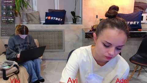 Media: Video of two women with light skin and brown hair in buns, working on laptops in a modern, well-lit office with a tiled floor, white walls, and plants.