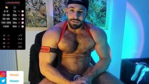 Media: Video of a muscular, bearded man with a red thong, wearing a black cap, sitting in a chair. Background includes a colorful wall art and a digital clock displaying 5:09, 70 degrees, and 100% humidity.