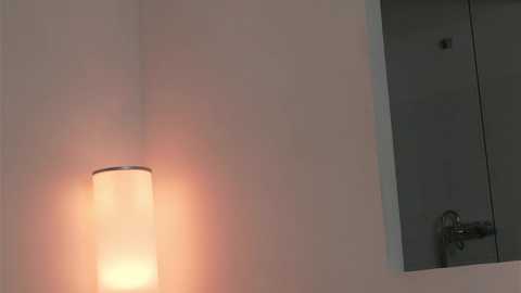 Media: Video of a minimalist bathroom interior featuring a modern, cylindrical, white-tiled lamp emitting a warm glow, mounted on a light beige wall. A sleek, silver faucet is partially visible on the right.