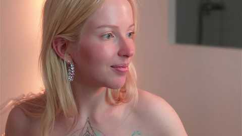 Media: Video of a blonde woman with fair skin and blue eyes, wearing a strapless dress and silver chandelier earrings, smiling gently, against a softly lit, plain beige wall background.