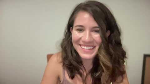 Media: Video of a young woman with wavy, dark brown hair, smiling, wearing a light-colored top, against a plain, light-colored wall background.