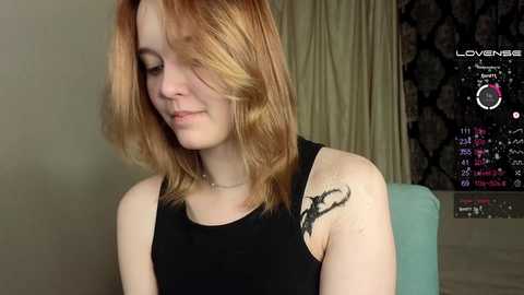 Media: Video of a fair-skinned woman with shoulder-length, wavy red hair, wearing a black sleeveless top, featuring a black tattoo of a bird on her left shoulder, in a dimly lit room with a dark, patterned wall and a TV screen displaying a calendar.