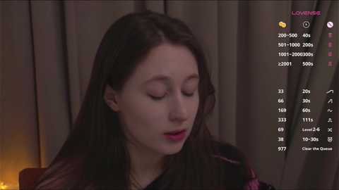 Video of a fair-skinned woman with long dark hair, wearing a black top, sitting with eyes closed, in a dimly lit room. A chat window with \"LOVINBRE\" and stats is visible.