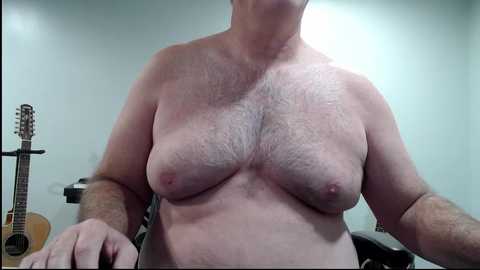 Media: Video of a topless, middle-aged, Caucasian man with pale skin, medium build, and average-sized breasts, standing indoors with a guitar and amplifier in the background.