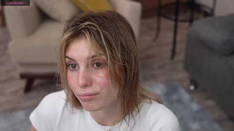 Media: Video of a young woman with messy, wet hair, wearing a white T-shirt, seated on a blue-patterned rug in a living room with beige furniture and a yellow blanket.