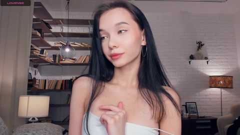 Video of a slender, fair-skinned woman with long black hair, wearing a white strapless top, standing in a modern loft with exposed brick walls, shelves of books, and a cozy lamp.