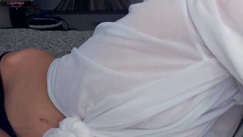 Media: A video of a person in a white, semi-transparent shirt, revealing their midriff and light-colored underwear, lying on a textured gray carpet in a dimly lit room.