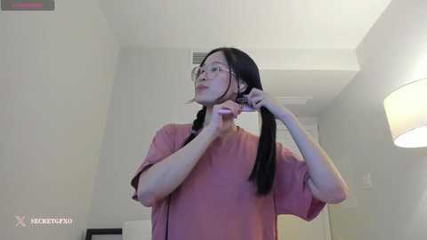 Media: A video of an Asian woman with long black hair, wearing glasses and a pink t-shirt, adjusting her hair in a modern, minimalist hotel room with white walls, a ceiling lamp, and a door in the background.