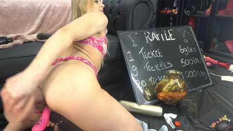 Media: A video of a blonde woman in pink lingerie, with a note on a blackboard detailing \"Tiffany,\" \"balix,\" \"tickle,\" \"fickles,\" \"5000x,\" and \"Pervy.\" The background includes a black couch, a red blanket, and scattered toys.