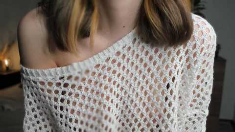 Media: Video of a woman with shoulder-length, multi-colored hair, wearing a white, off-shoulder crochet sweater, revealing cleavage. Background shows a dimly lit room with a lit candle and a blurred, dark wooden dresser.