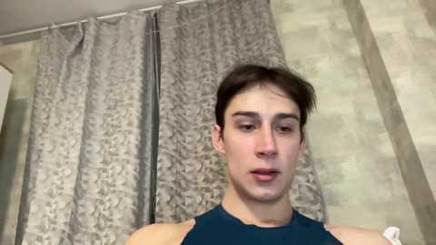 Media: Video of a young Caucasian man with short brown hair, wearing a dark blue sleeveless shirt, standing in a bathroom with gray patterned shower curtain and beige tiled walls.