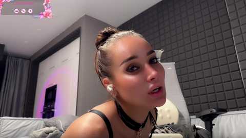 Video of a young woman with light skin, brown hair in a bun, wearing a black choker and white pearls, posing on a bed with grey walls and a grey quilted headboard.