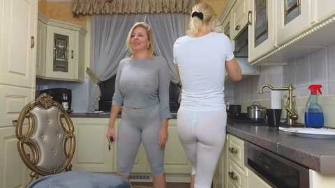 Media: Video of two blonde women in matching grey and white workout gear, smiling, in a modern kitchen with cream cabinets and beige walls.