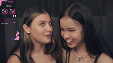 Media: Video of two smiling young women with light skin and long dark hair, wearing casual outfits, indoors.