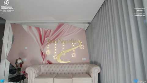 Media: Video of a modern, luxurious room with a large, pink floral mural on the wall. A beige tufted sofa is centered with a gold harp-shaped light fixture above. Grey vertical blinds cover the windows on the right.