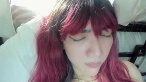 Media: Video of a young woman with long, straight, pink and black hair, lying on a white pillow, eyes closed, mouth slightly open, wearing a gold necklace.