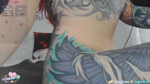 Media: A video of a woman with extensive, colorful tattoos on her back, showcasing a blue feather design, lying on a bed with a gray blanket and a red pillow.