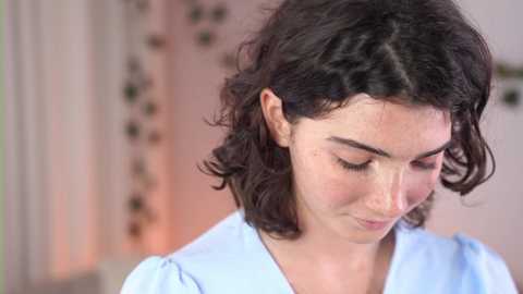 Media: Video of a young woman with shoulder-length, curly dark hair, wearing a light blue blouse, looking down, with a soft, blurred background featuring floral wallpaper.