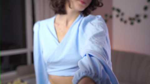 Media: A video of a woman with short, curly brown hair, wearing a light blue, short-sleeved top, taken indoors with a blurred background.