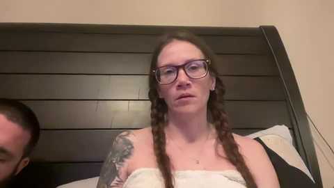 Media: Video of a Caucasian woman with long brown hair in braids, wearing glasses and a white towel, with a large tattoo on her left arm, standing in front of a dark wooden bed frame.