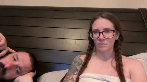 Media: Video of a pale-skinned woman with glasses and braided hair, wearing a towel, and a man with a beard, lying on a dark wooden headboard in a bedroom.