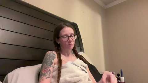 Media: Video of a fair-skinned woman with long brown braids, glasses, and tattoos, sitting on a bed with beige walls and a wooden headboard, wrapped in a light blanket, holding a phone.