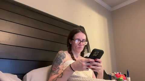 Media: Video of a Caucasian woman with glasses, tattoos, and braided hair, sitting on a bed in a beige room, looking at a smartphone.