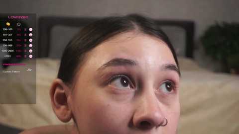 Media: A close-up video of a young Asian woman with dark hair, large eyes, and a septum piercing, seen from the side. The background shows a bed with beige sheets and a potted plant.