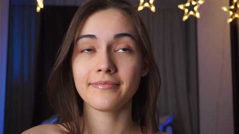 Media: Video of a young woman with fair skin, straight brown hair, and light makeup, smiling gently. She wears a light-colored top. Background features dark curtains and warm, glowing star-shaped lights.