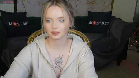 Media: Video of a young blonde woman with fair skin and blue eyes, wearing a beige hoodie, seated on a dark sofa with \"MSBET\" pillows, against a floral wallpaper backdrop.