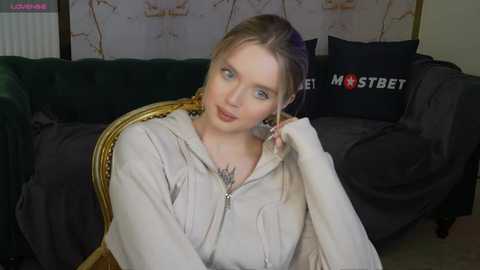 Media: Video of a young, fair-skinned woman with blonde hair, wearing a beige hoodie, sitting on a dark green couch. Background features a black pillow with white text, and a beige wall with abstract art.