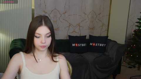 Media: Video of a young Caucasian woman with straight brown hair, fair skin, and a slender physique, wearing a white tank top. She sits on a dark green couch with a black pillow featuring \"Masters of the Universe\" text.