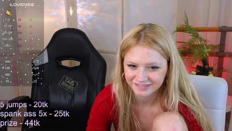 Media: Video of a young, fair-skinned woman with long blonde hair, wearing a red top, sitting in a black gaming chair. Background includes a calendar, potted plant, and a white cushion. Text overlays mention a Twitch stream with 25k views and 4.4k prize.