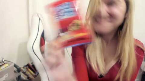 Media: A video of a smiling young woman with long blonde hair, wearing a red top, holding a white dress with a red box. Background shows a white wall and a cluttered desk with books and a lamp.