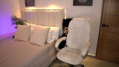 Media: Video of a modern bedroom with a white tufted headboard, plush pillows, beige bedding, a wooden door, and a white ergonomic office chair with a black teddy bear on the seat.