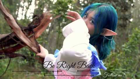 Media: Video of a blue-haired, elf-like cosplay character with pointy ears, wearing a white turtleneck and blue skirt, reaching for a branch in a forest. Text reads: \"Be Right Back.\