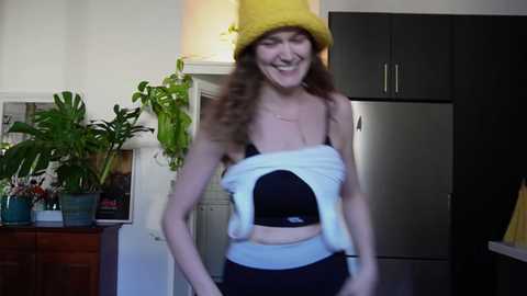 Media: A video of a joyful, smiling woman with curly brown hair, wearing a yellow hat, black bra, and white pants, in a modern kitchen with green plants and stainless steel appliances.